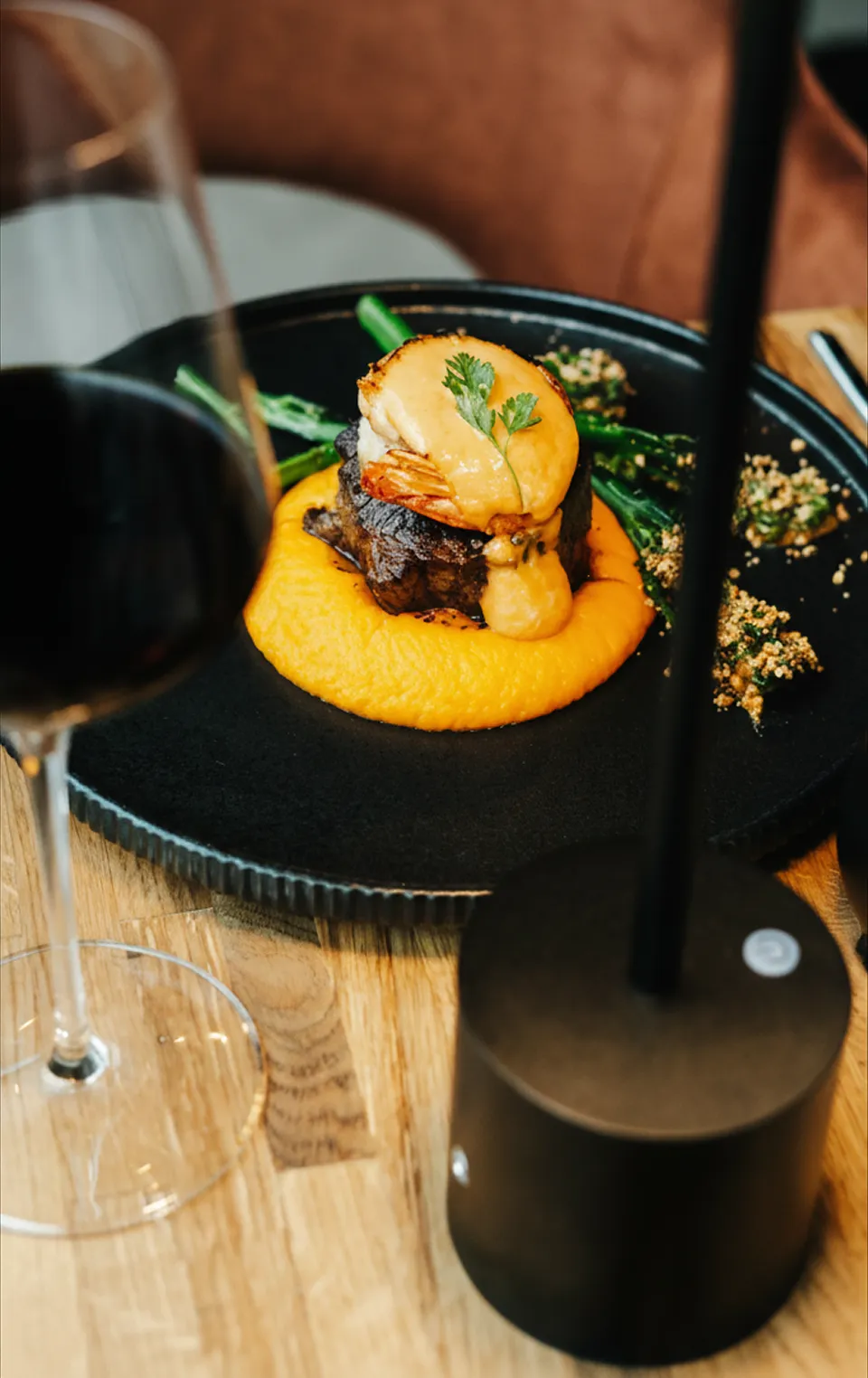 Gourmet dish with steak, shrimp topped with sauce and greens served on orange puree on a black plate.